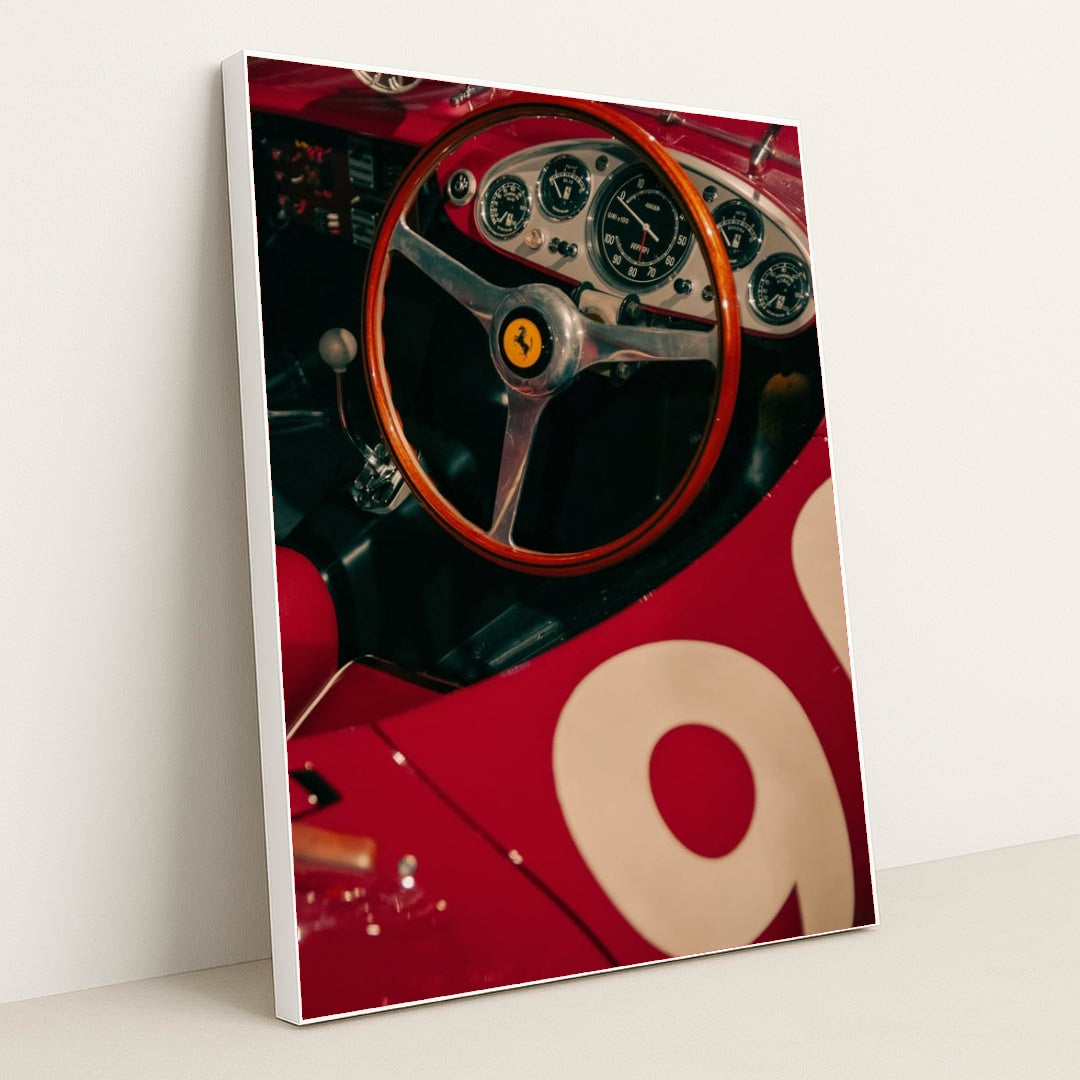 This photo shows the cockpit of a classic red Ferrari race car with a wooden steering wheel and vintage gauges, in a white frame.