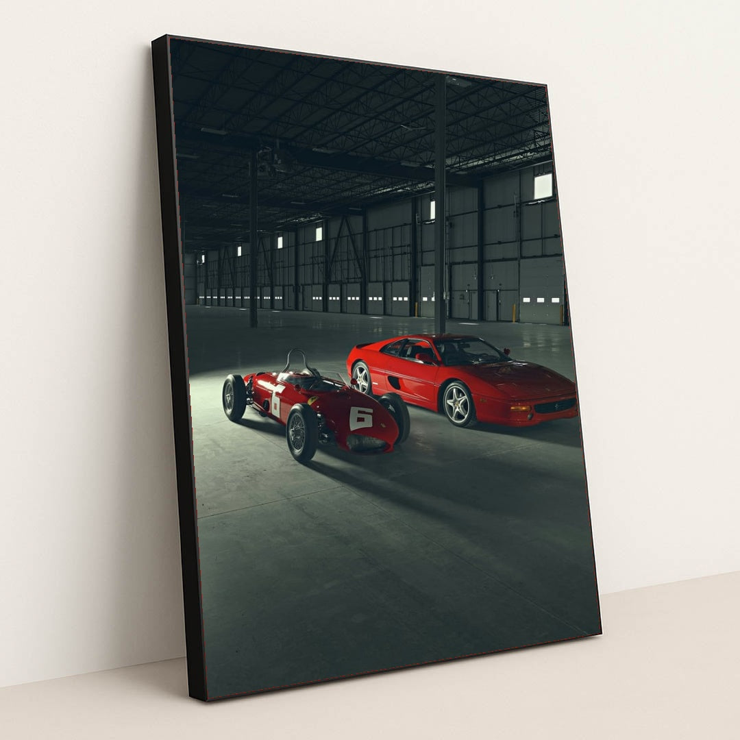 This photo shows a vintage red Ferrari race car and a red Ferrari coupe parked inside a large industrial warehouse, in a black frame.
