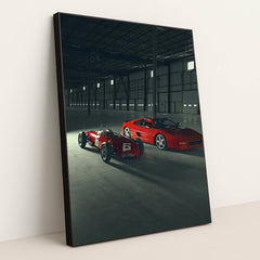 This photo shows a vintage red Ferrari race car and a red Ferrari coupe parked inside a large industrial warehouse, in a black frame.