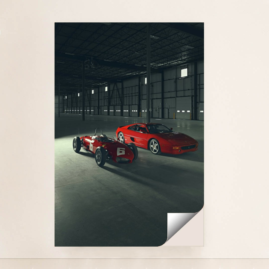 This photo shows a vintage red Ferrari race car and a red Ferrari coupe parked inside a large industrial warehouse, minimalist unframed style.