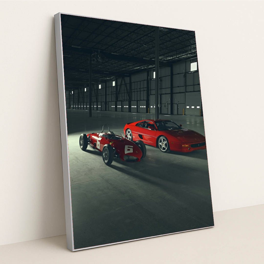 This photo shows a vintage red Ferrari race car and a red Ferrari coupe parked inside a large industrial warehouse, in a silver frame.