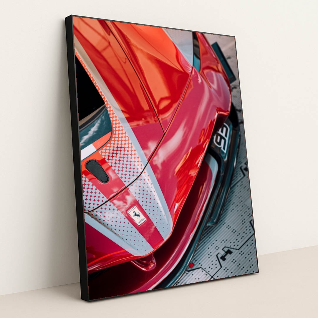 This photo shows a close-up view of a red Ferrari with dotted racing stripes reflecting dramatic skies, in a black frame.