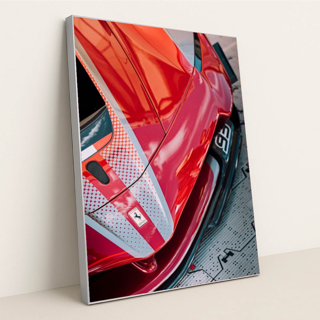 This photo shows a close-up view of a red Ferrari with dotted racing stripes reflecting dramatic skies, in a silver frame.