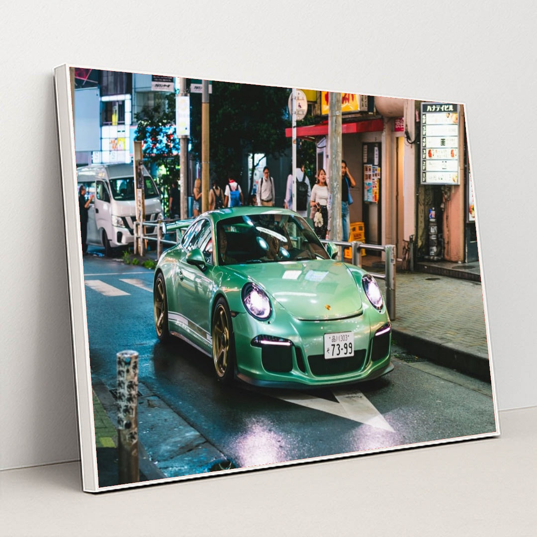 This photo shows a mint green Porsche sports car with gold wheels driving through a vibrant city street at night, in a white frame.