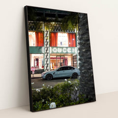 This photo shows a white BMW sedan parked in front of an illuminated Gucci store at night, in a black frame.