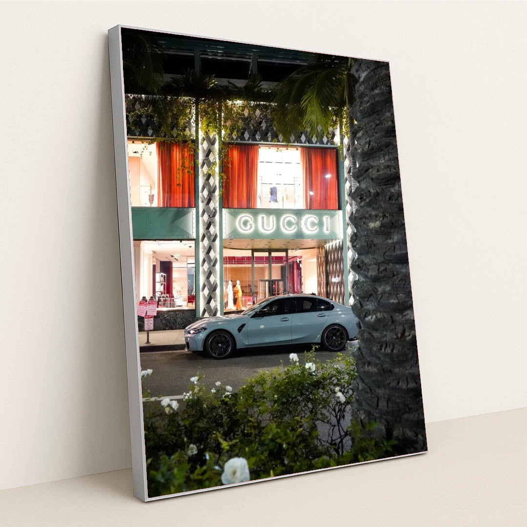 This photo shows a white BMW sedan parked in front of an illuminated Gucci store at night, in a silver frame.