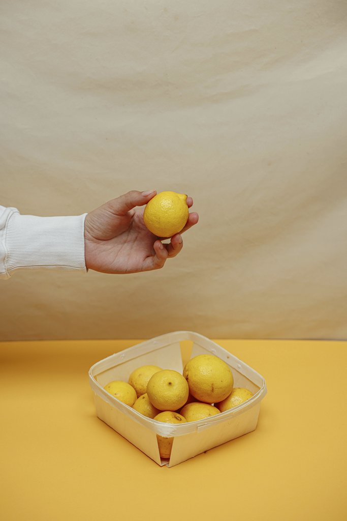Cup of  Couple - A hand holds a lemon above a basket filled with lemons on a yellow surface, set against a plain backdrop.