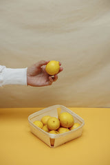 Cup of  Couple - A hand holds a lemon above a basket filled with lemons on a yellow surface, set against a plain backdrop.