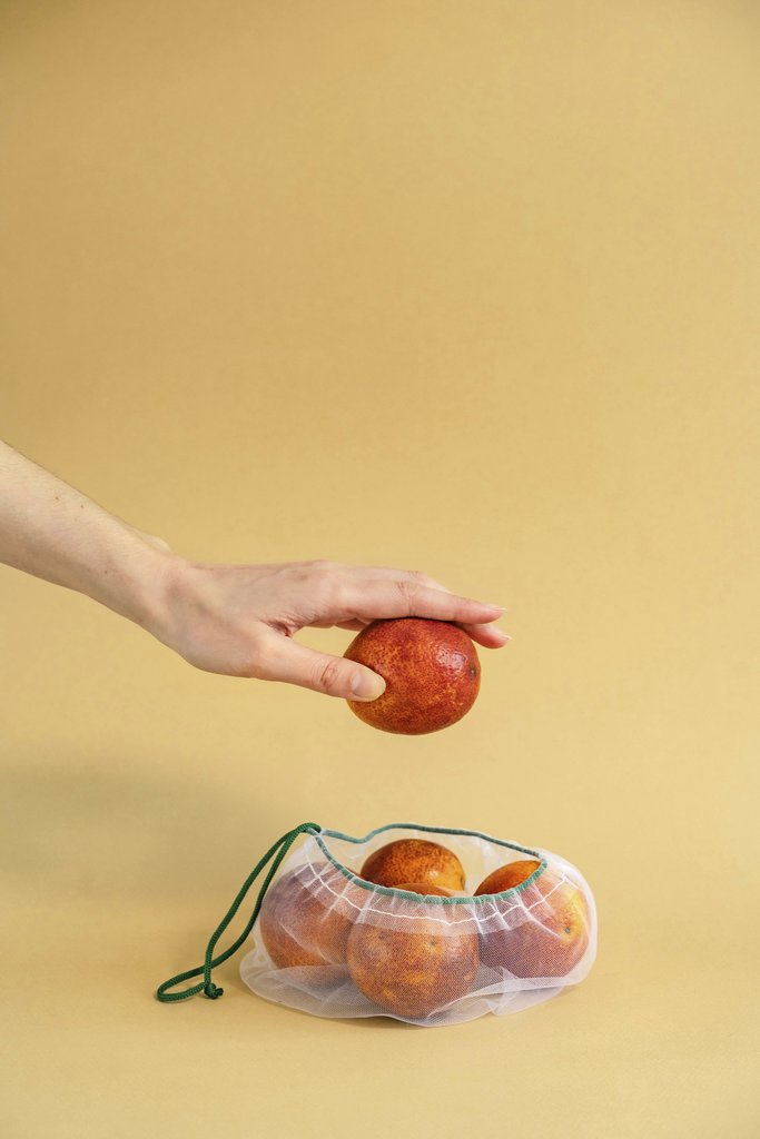 Mikhail Nilov - A hand reaches for a ripe peach in a reusable mesh bag, set against a yellow backdrop.