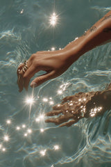 This artwork features a hand with rings reaching into sparkling blue water.