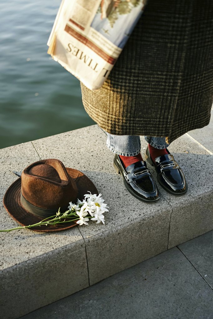 Beyzanur K. - Elegant shoes near water, with a hat, newspaper, and flowers on a sunny day.
