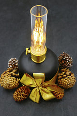 Marta Branco - A stylish black lamp with glowing filament surrounded by pine cones and adorned with a golden ribbon, perfect for festive decor.
