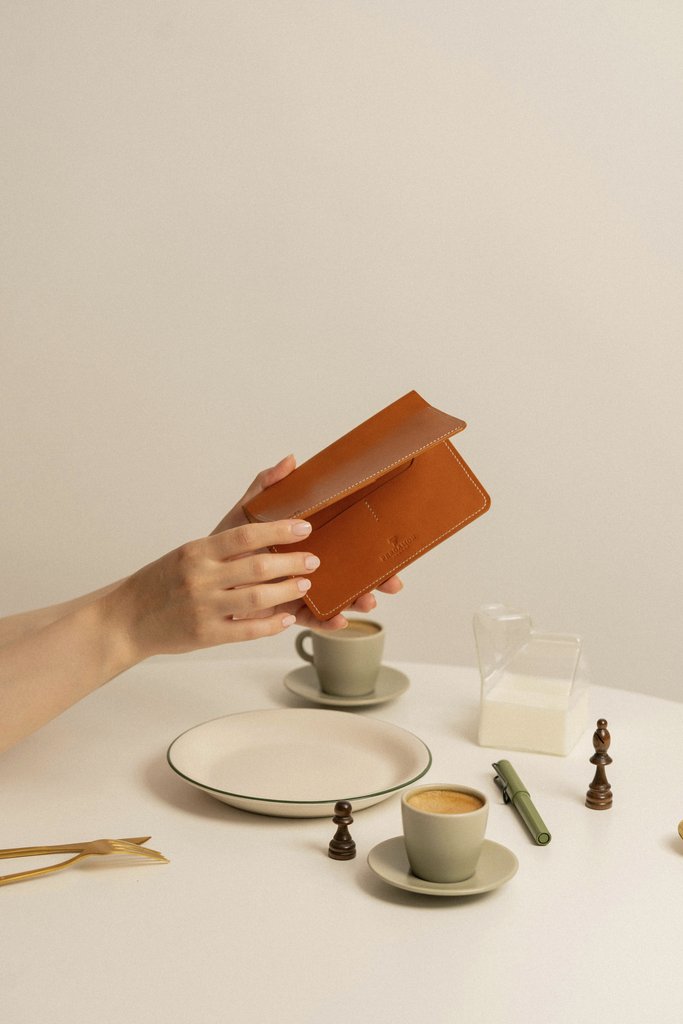 Gül Işık - A close-up of a leather wallet held over a café table with coffee, chess pieces, and minimalistic decor.