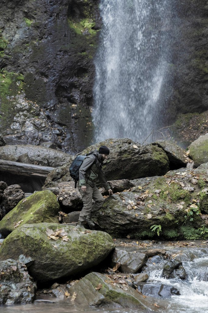 Saleh  Bakhshiyev - Adventurer climbs mossy rocks by a waterfall in a lush forest setting.