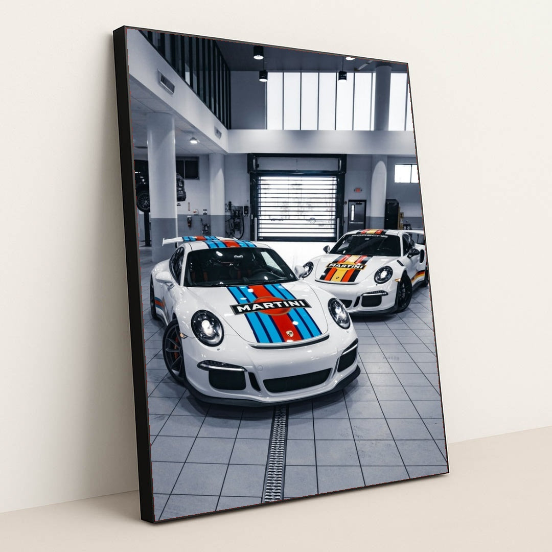 This photo shows two Porsche sports cars with Martini Racing liveries parked in a bright modern garage, in a black frame.