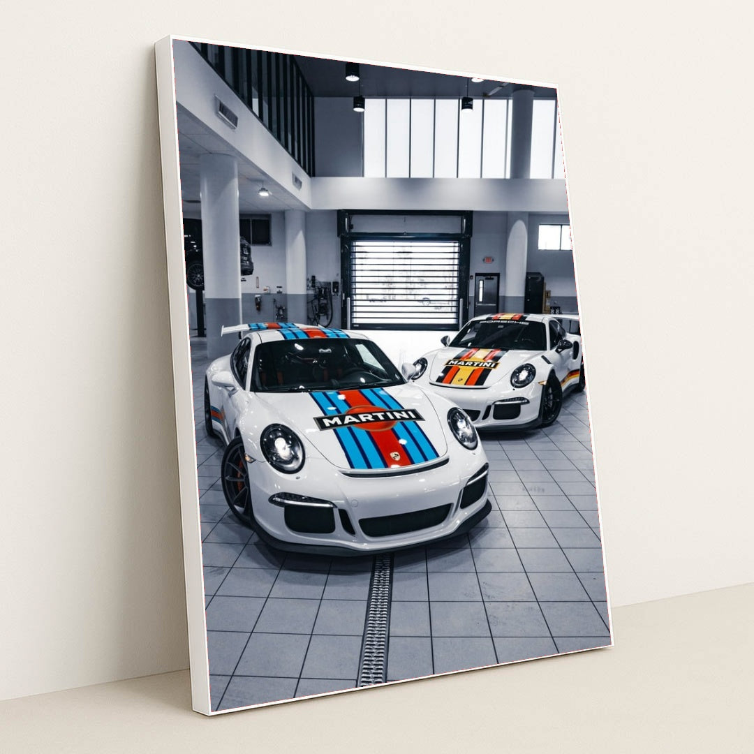 This photo shows two Porsche sports cars with Martini Racing liveries parked in a bright modern garage, in a white frame.