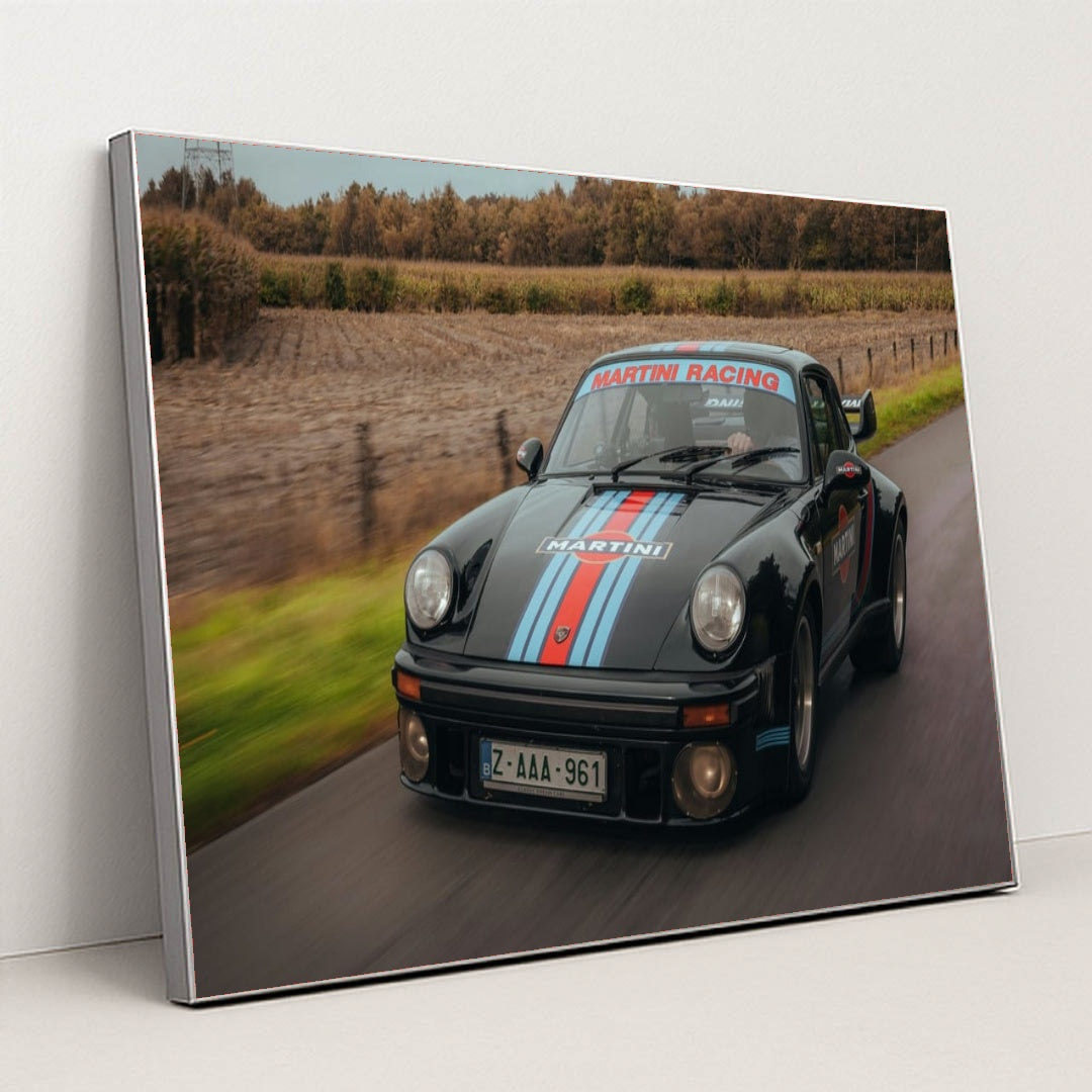 This photo features a vintage black Porsche with Martini Racing stripes driving on a rural road, in a silver frame.