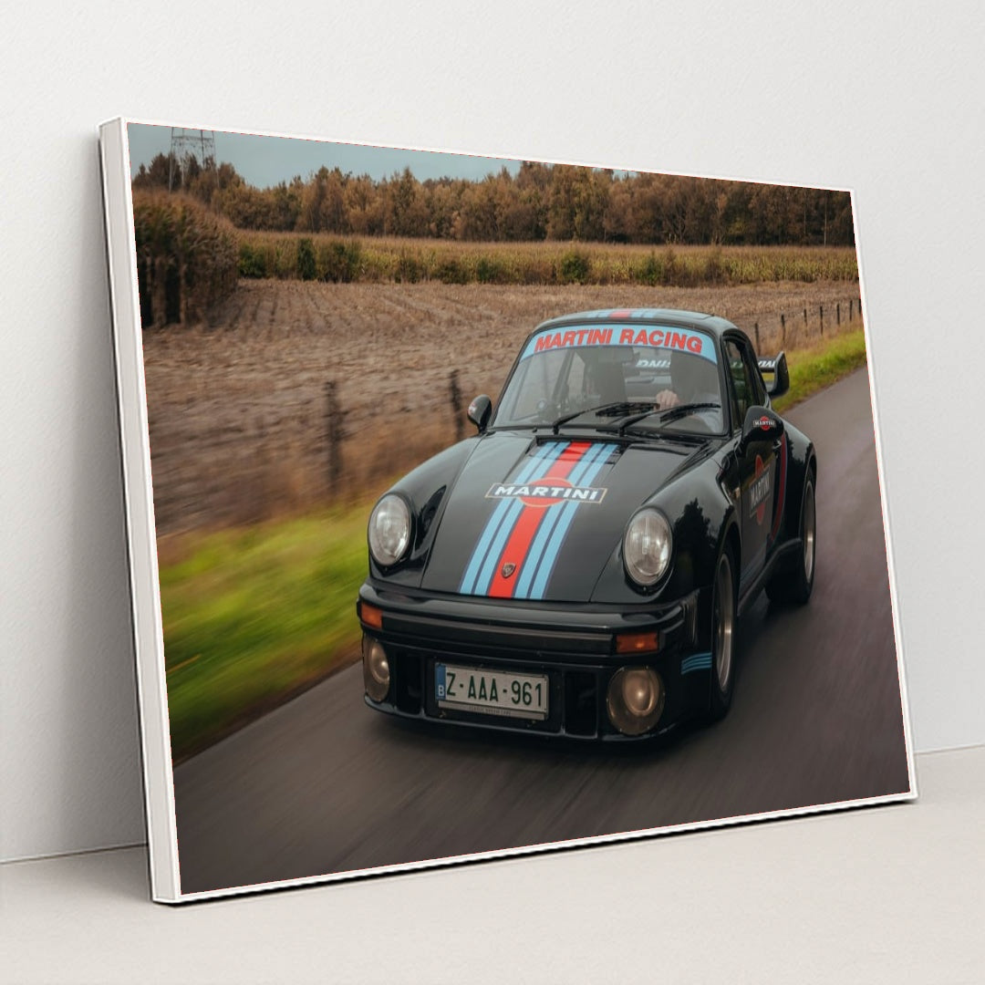 This photo features a vintage black Porsche with Martini Racing stripes driving on a rural road, in a white frame.