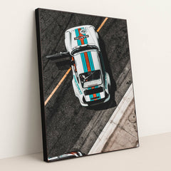 This photo shows an overhead view of a white Porsche race car with Martini stripes and an open door on a racetrack, in a black frame.