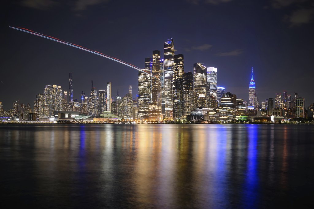 Attie Heunis - Stunning nighttime view of New York City skyline across the reflective waterfront, featuring illuminated skyscrapers.