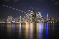 Attie Heunis - Stunning nighttime view of New York City skyline across the reflective waterfront, featuring illuminated skyscrapers.