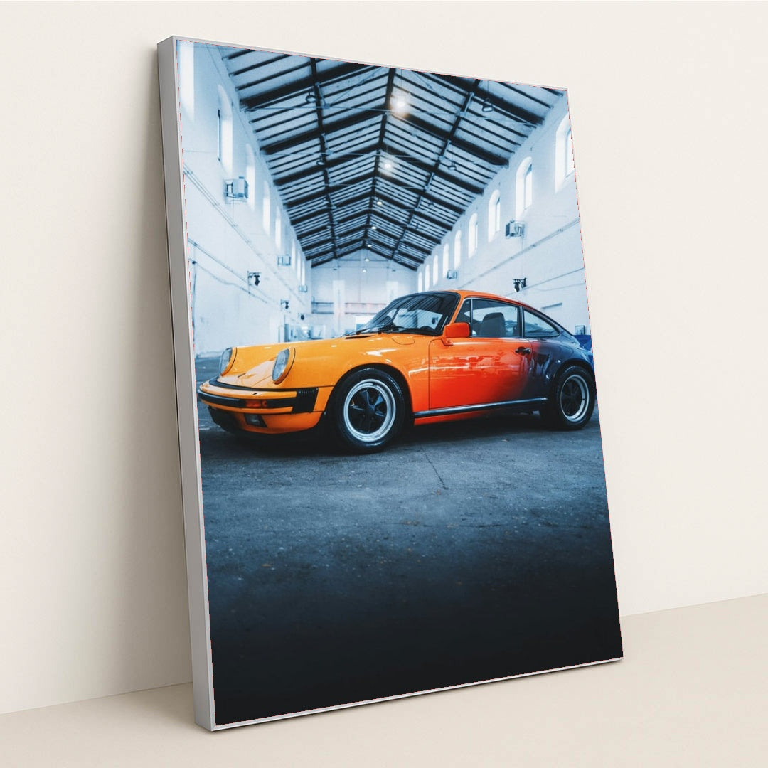 This photo features an orange and red classic Porsche sports car parked inside a tall industrial warehouse, in a silver frame.