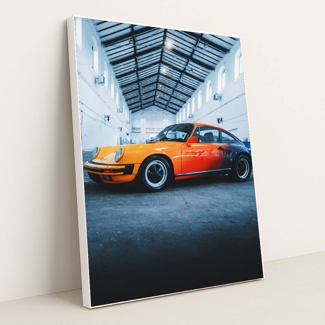 This photo features an orange and red classic Porsche sports car parked inside a tall industrial warehouse, in a white frame.