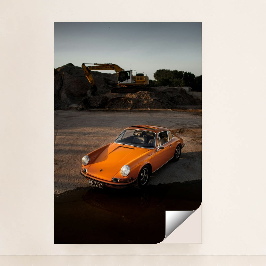 This photo shows an orange classic Porsche 911 parked on gravel at sunset with a yellow excavator in the background, minimalist unframed style.