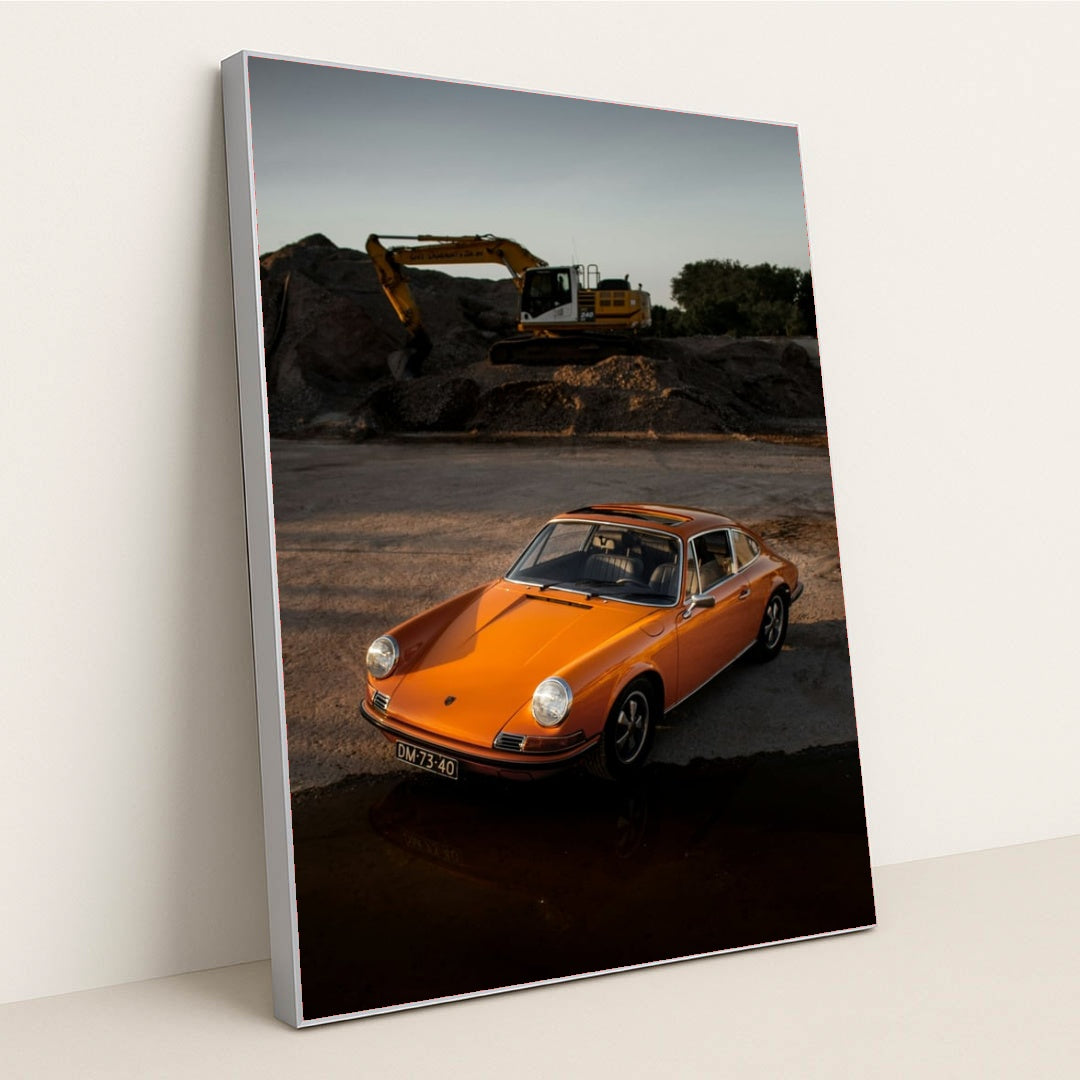 This photo shows an orange classic Porsche 911 parked on gravel at sunset with a yellow excavator in the background, in a silver frame.
