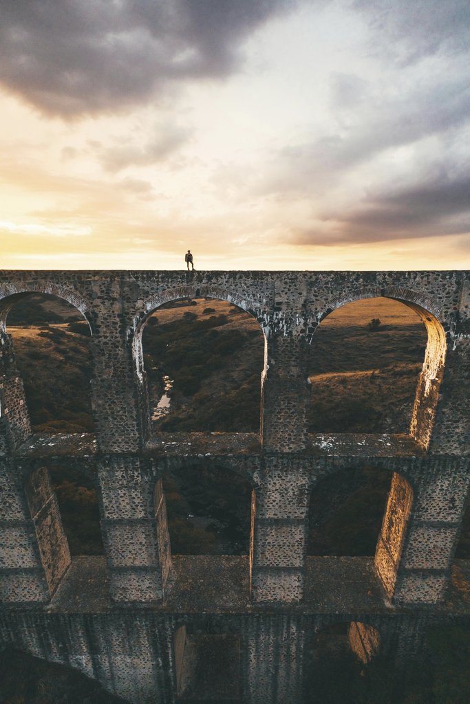 Mikhail Nilov - A lone figure stands atop a historic aqueduct at sunset, surrounded by dramatic clouds.
