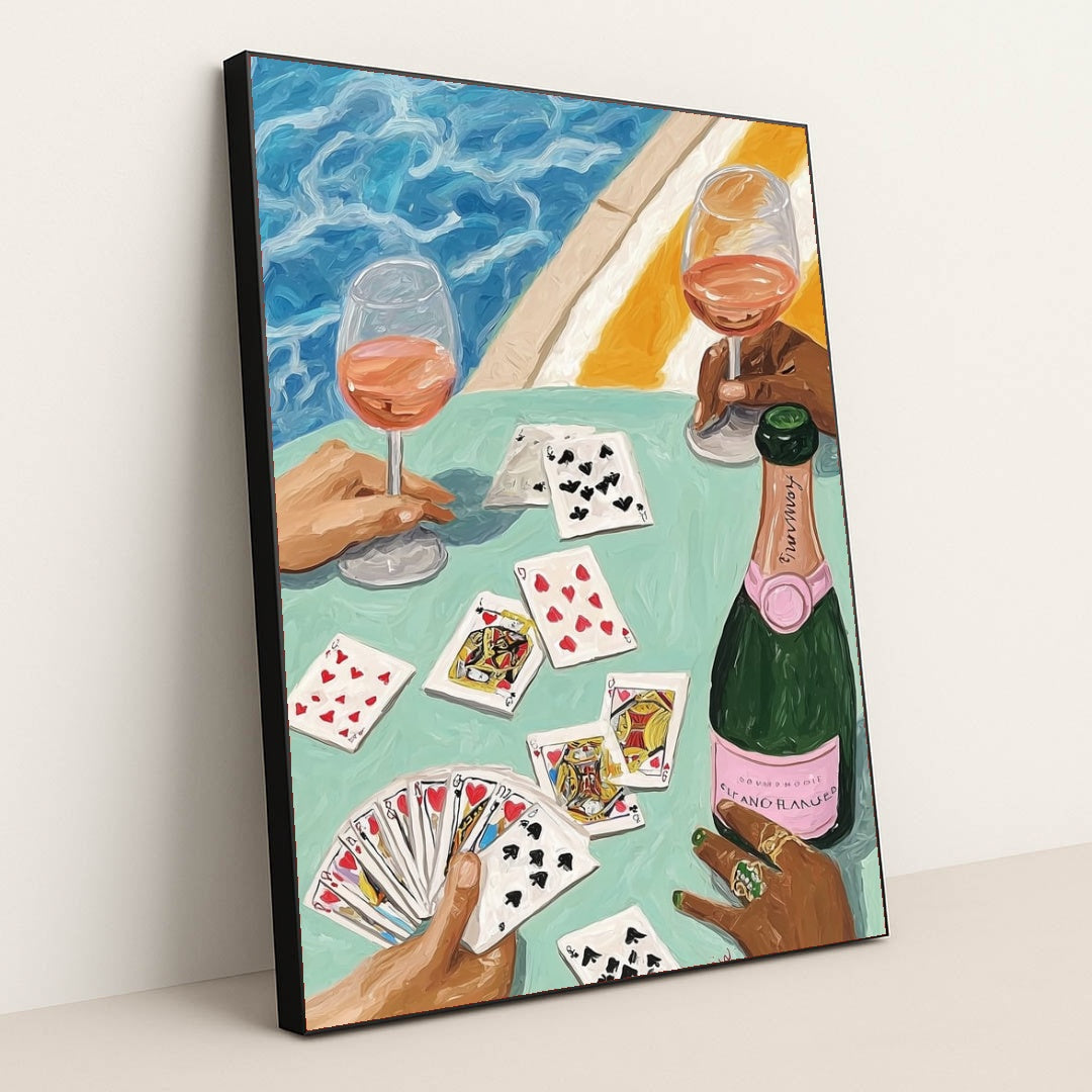 This artwork features two hands holding wine glasses, playing cards, and a bottle of rosé by a pool, in a black frame.