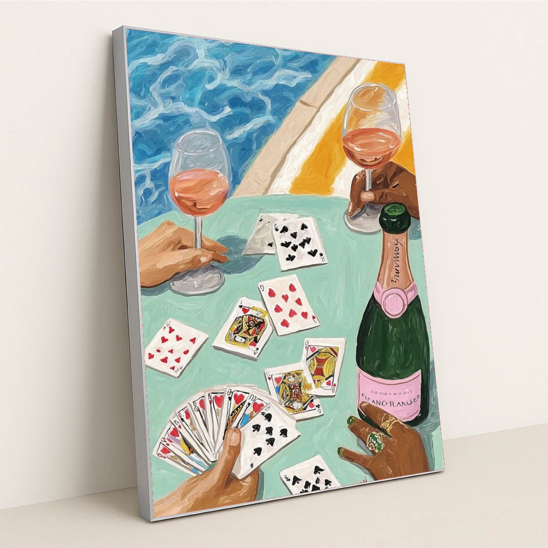 This artwork features two hands holding wine glasses, playing cards, and a bottle of rosé by a pool, in a silver frame.