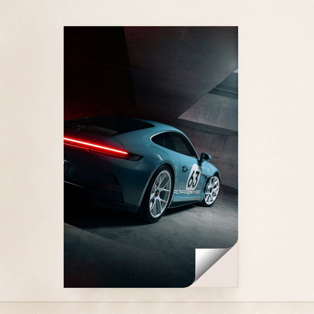 This photo shows a silver Porsche sports car with number 63 and glowing taillights in a modern concrete garage, unframed.