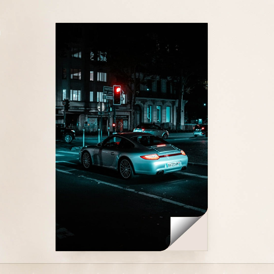 This photo shows a silver Porsche 911 coupe waiting at a red traffic light on a city street at night, minimalist unframed style.