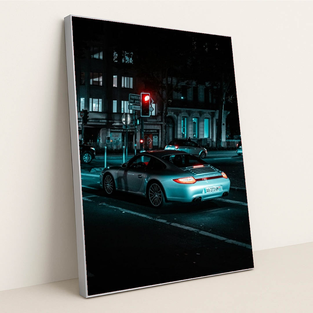 This photo shows a silver Porsche 911 coupe waiting at a red traffic light on a city street at night, in a silver frame.