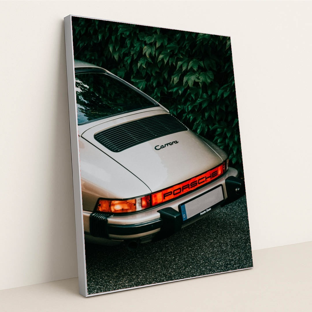 This photo shows the rear end of a classic silver Porsche Carrera parked in front of green leafy foliage, in a silver frame.