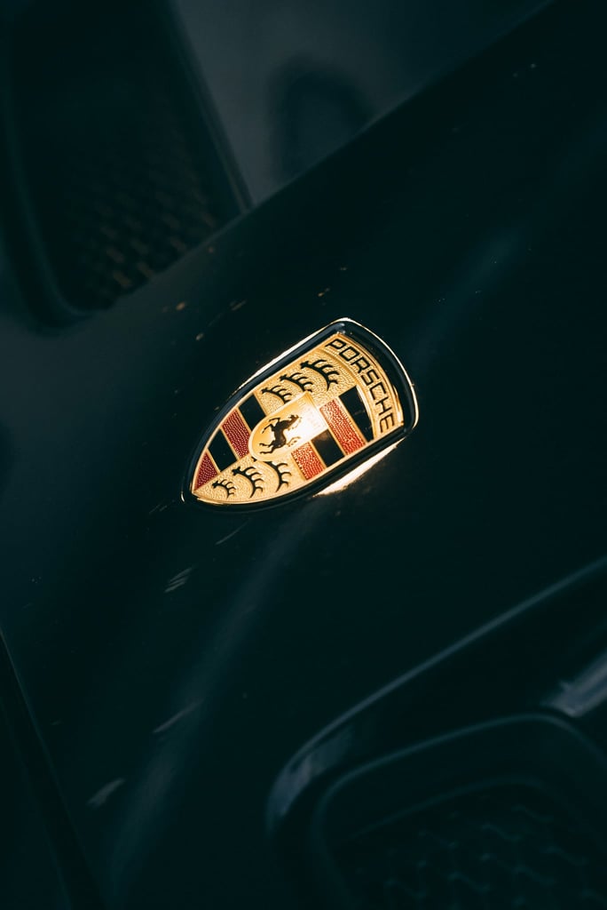 Close-up of the Porsche Emblem in Focus on a black hood, showcasing its gold and crimson details as car wall art.
