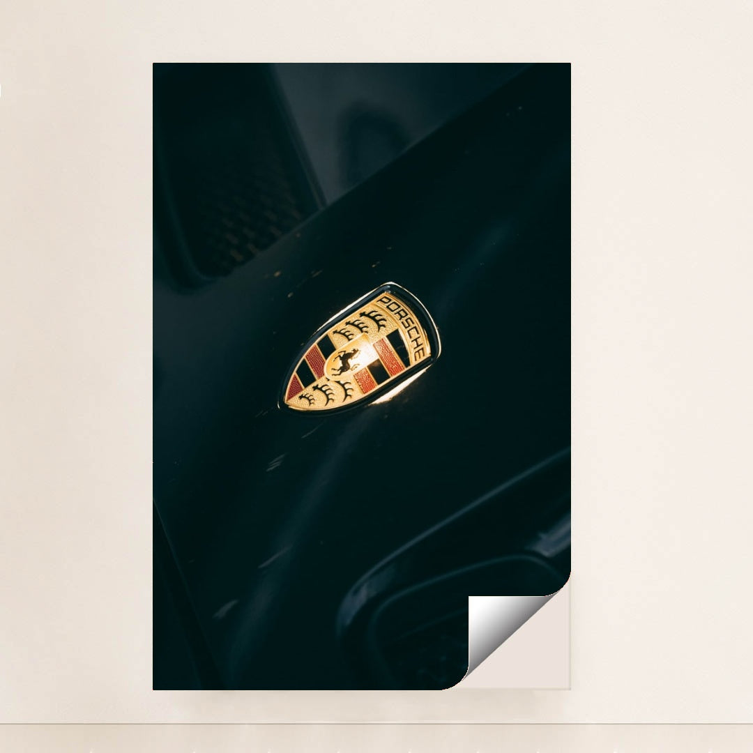 This photo shows a close-up of a classic gold and black Porsche crest badge on a dark car hood, unframed.