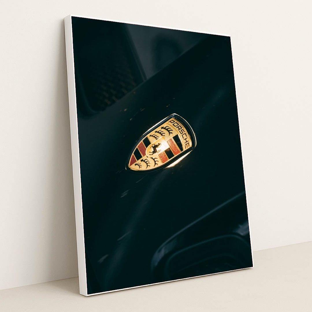 This photo shows a close-up of a classic gold and black Porsche crest badge on a dark car hood, in a white frame.