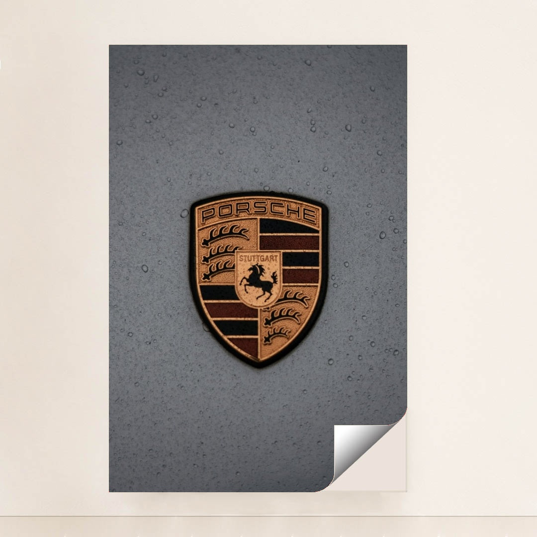 This photo features a gold Porsche emblem with raindrops on a gray car hood, unframed.