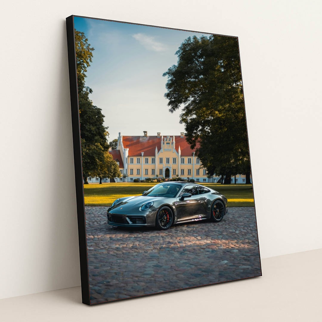 This photo shows a silver Porsche sports car parked on a cobblestone driveway in front of a large historic mansion, in a black frame.