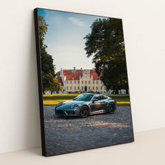 This photo shows a silver Porsche sports car parked on a cobblestone driveway in front of a large historic mansion, in a black frame.