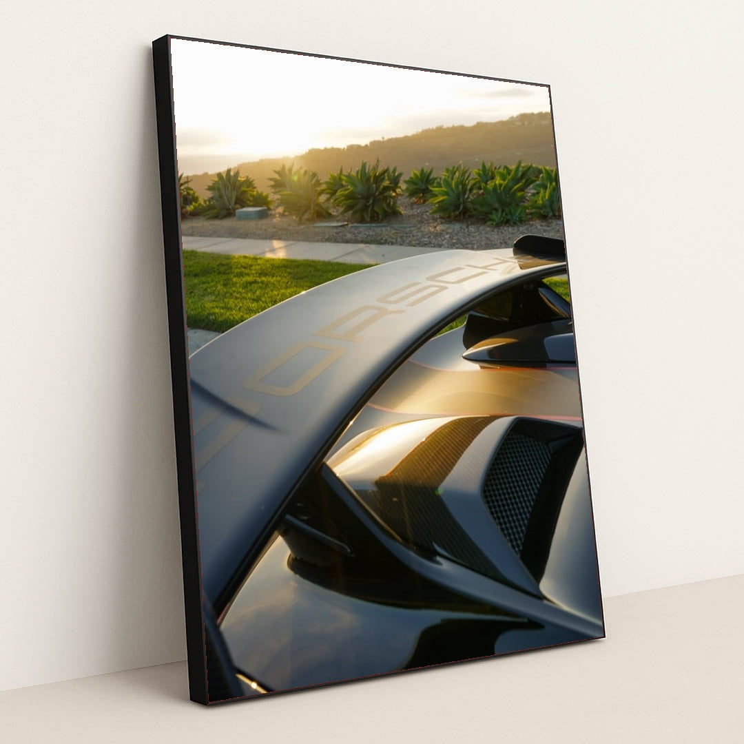 This photo shows a Porsche GT wing and rear vents at sunset with greenery and hills in the background, in a black frame.