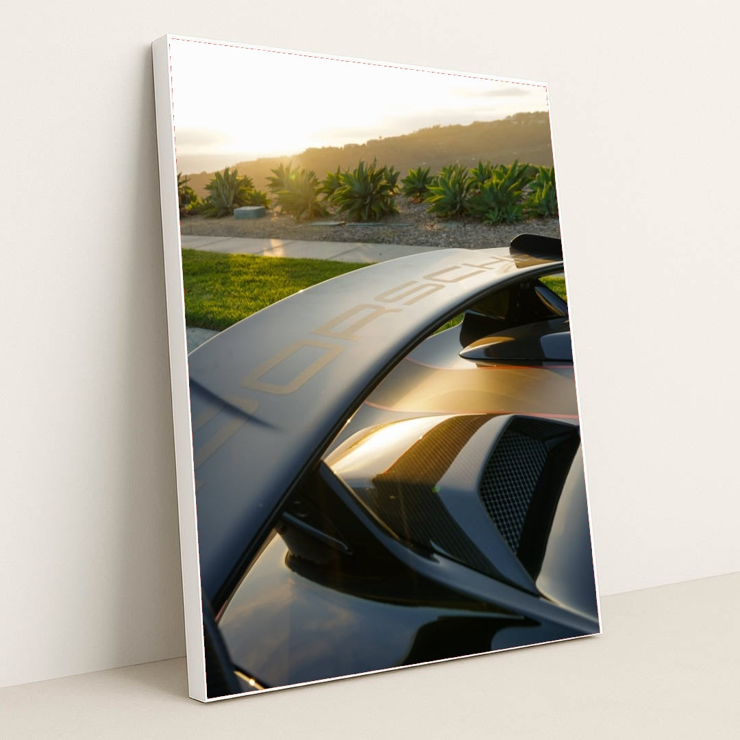 This photo shows a Porsche GT wing and rear vents at sunset with greenery and hills in the background, in a white frame.