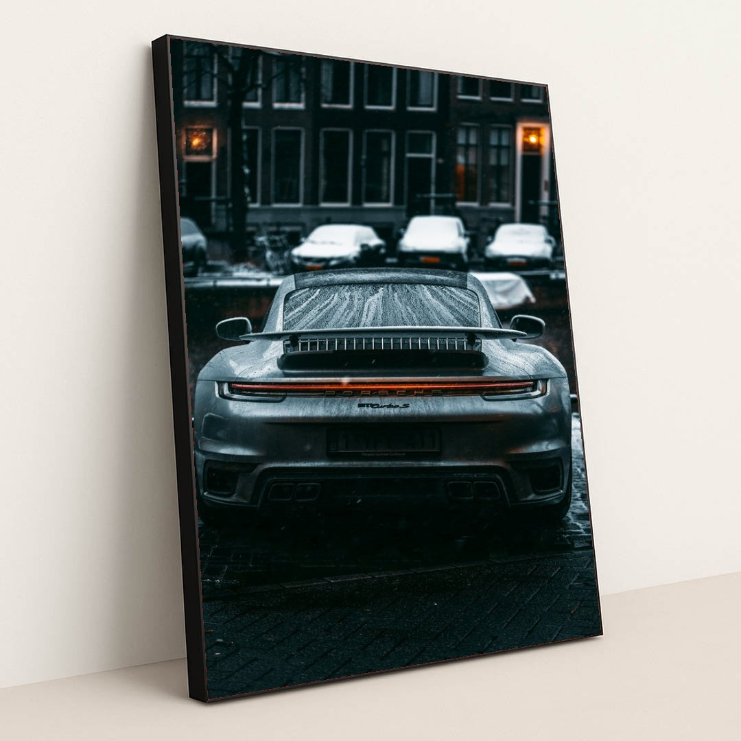 This photo shows the rear view of a silver Porsche sports car covered in raindrops on a city street at night, in a black frame.