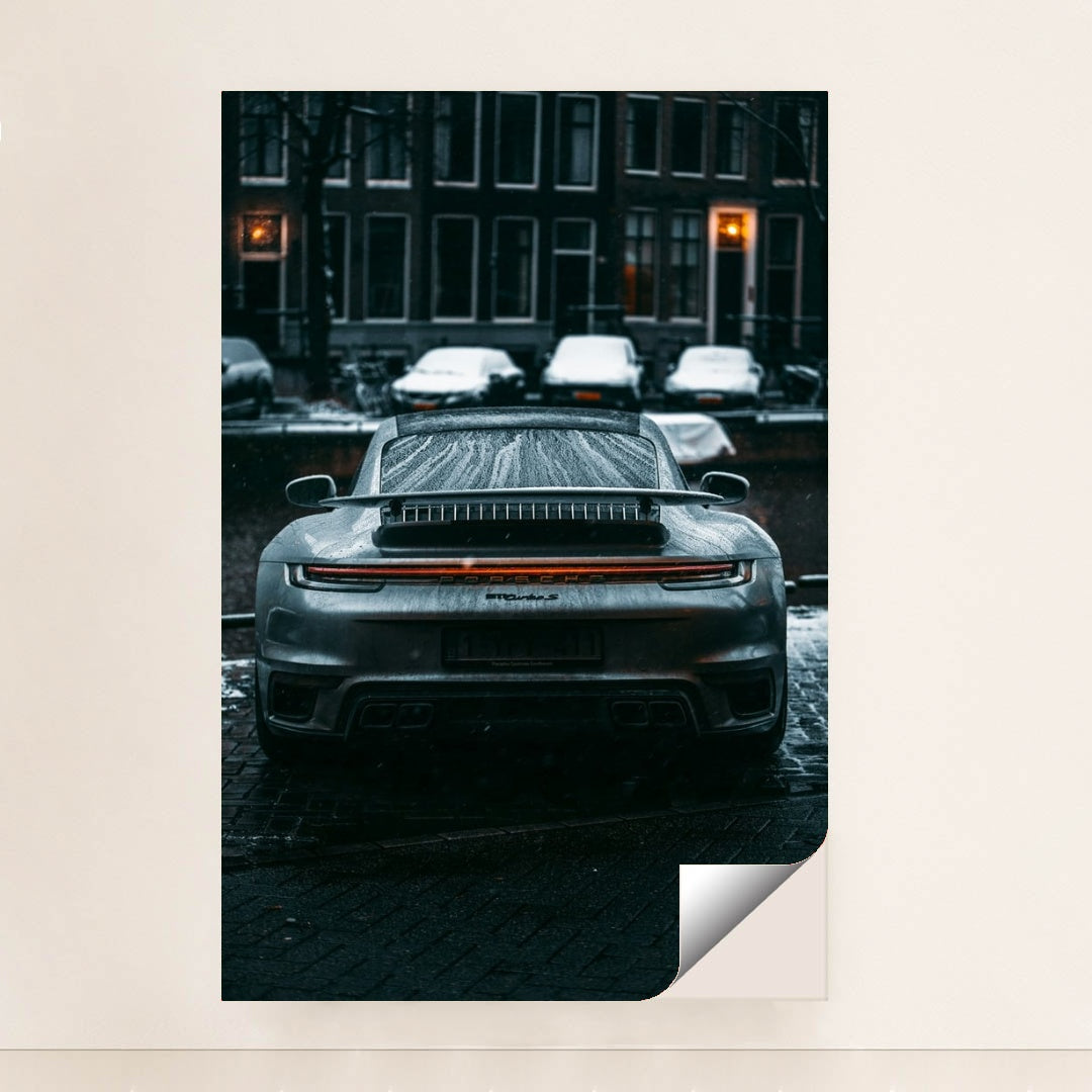 This photo shows the rear view of a silver Porsche sports car covered in raindrops on a city street at night, unframed.