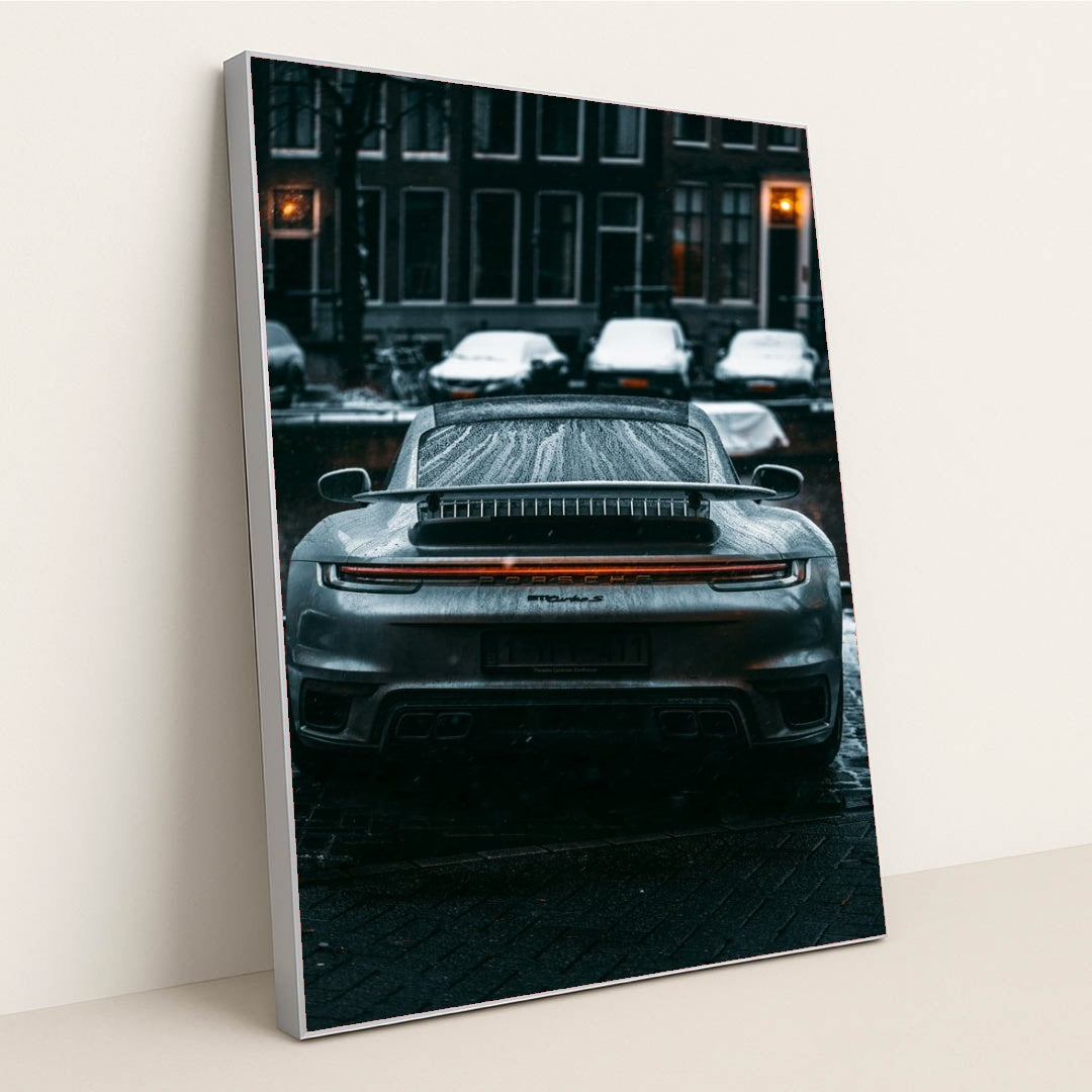 This photo shows the rear view of a silver Porsche sports car covered in raindrops on a city street at night, in a silver frame.