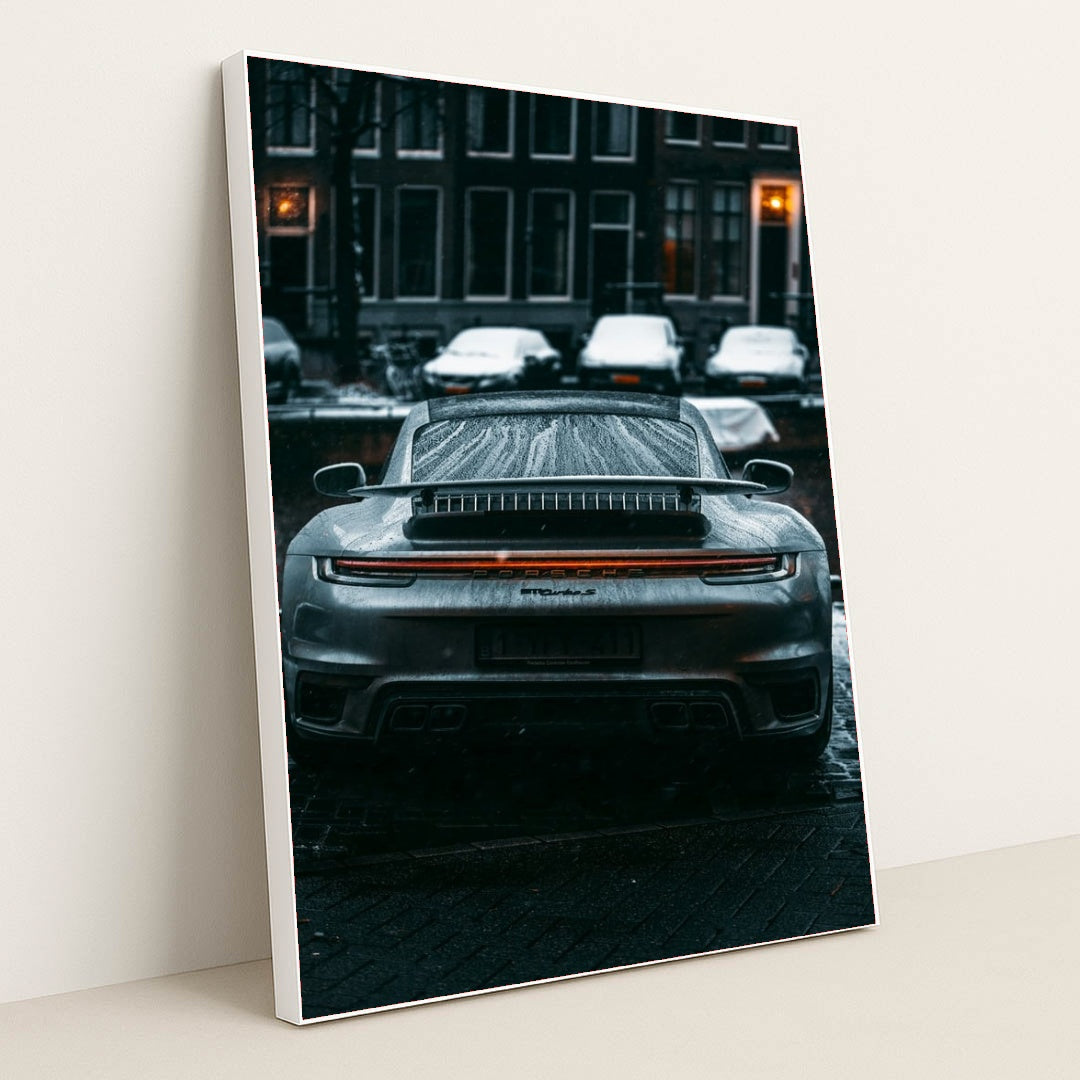 This photo shows the rear view of a silver Porsche sports car covered in raindrops on a city street at night, in a white frame.