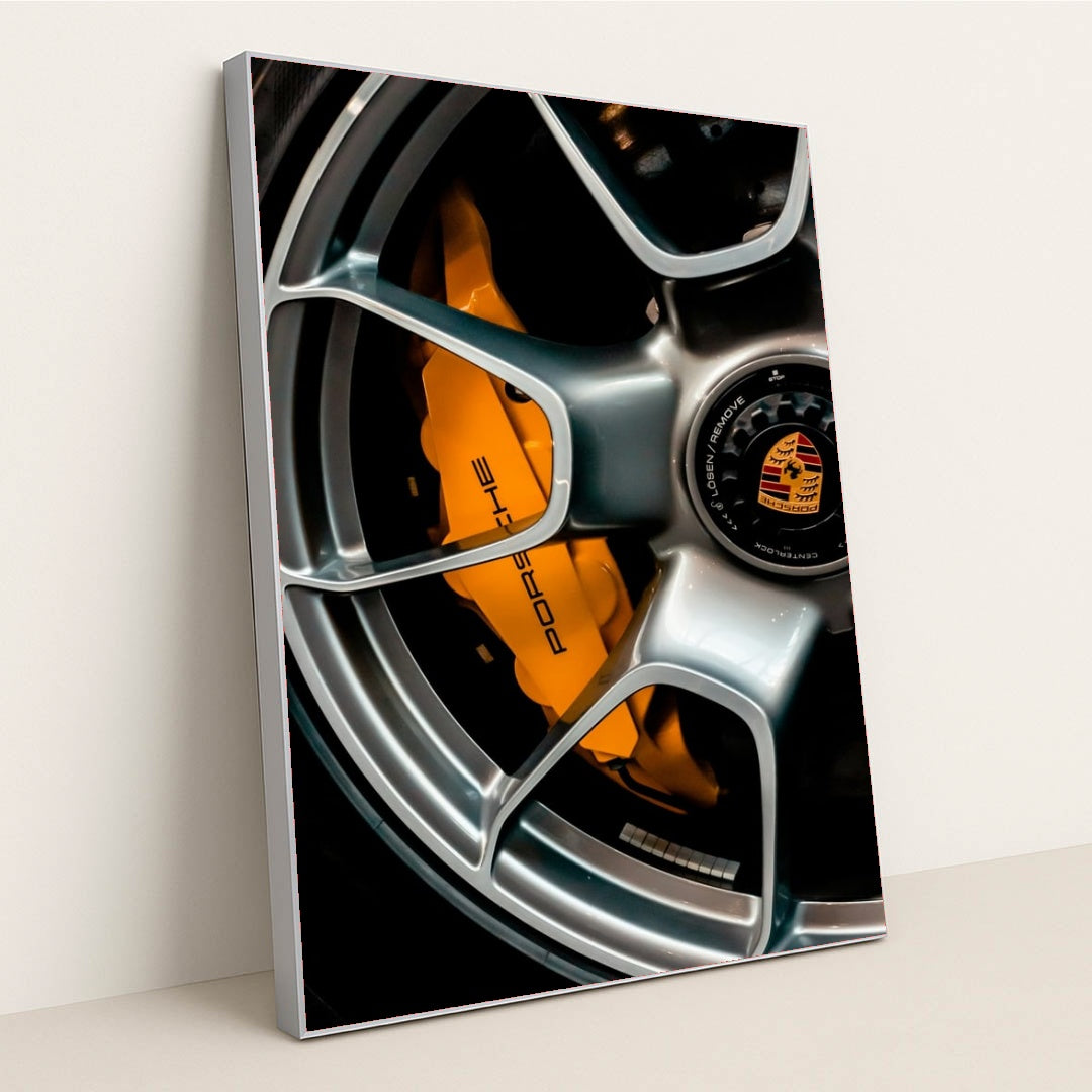 This photo shows a close-up of a silver Porsche wheel with an orange brake caliper and crest logo, in a silver frame.