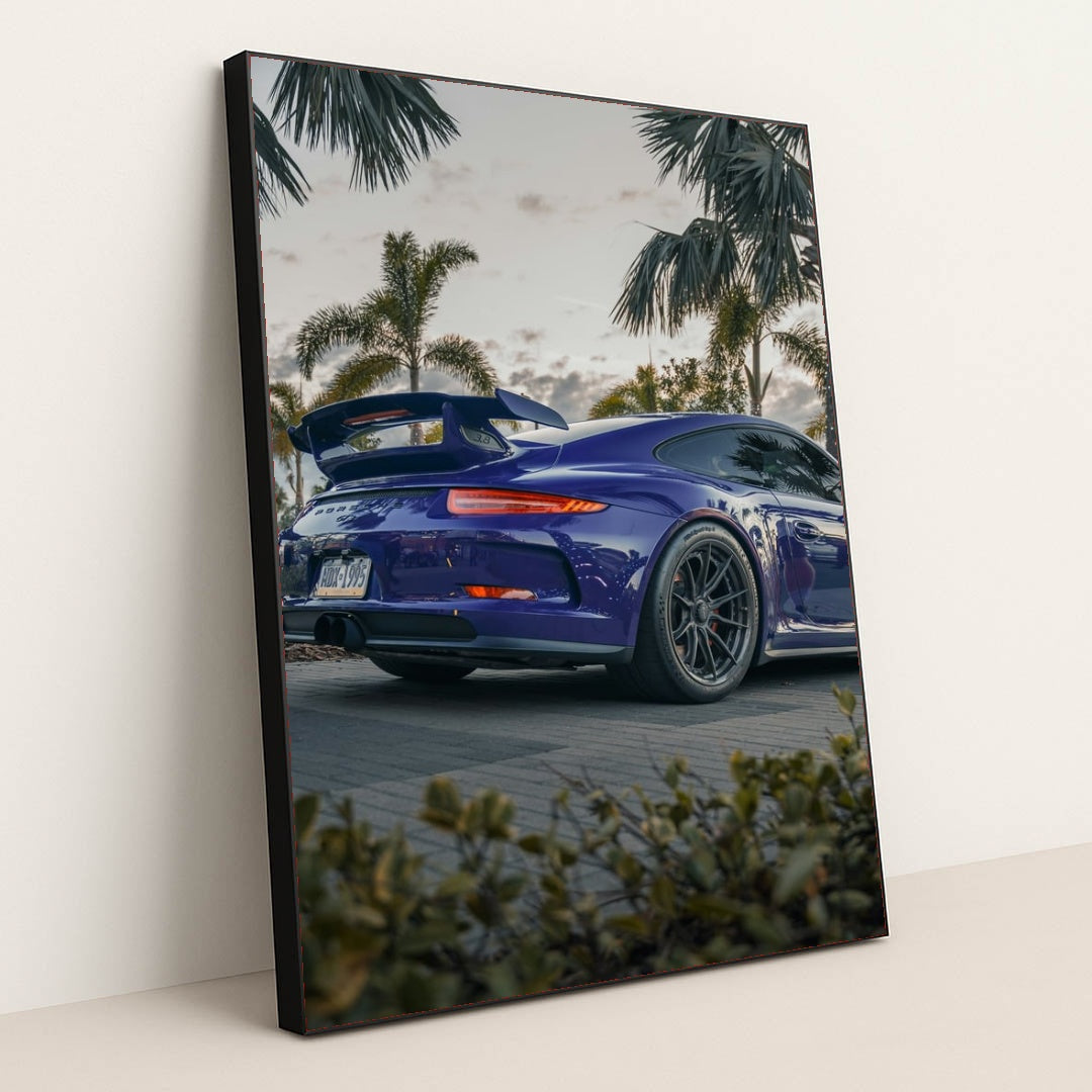 This photo shows a blue Porsche sports car with a large rear wing parked among palm trees at dusk, in a black frame.
